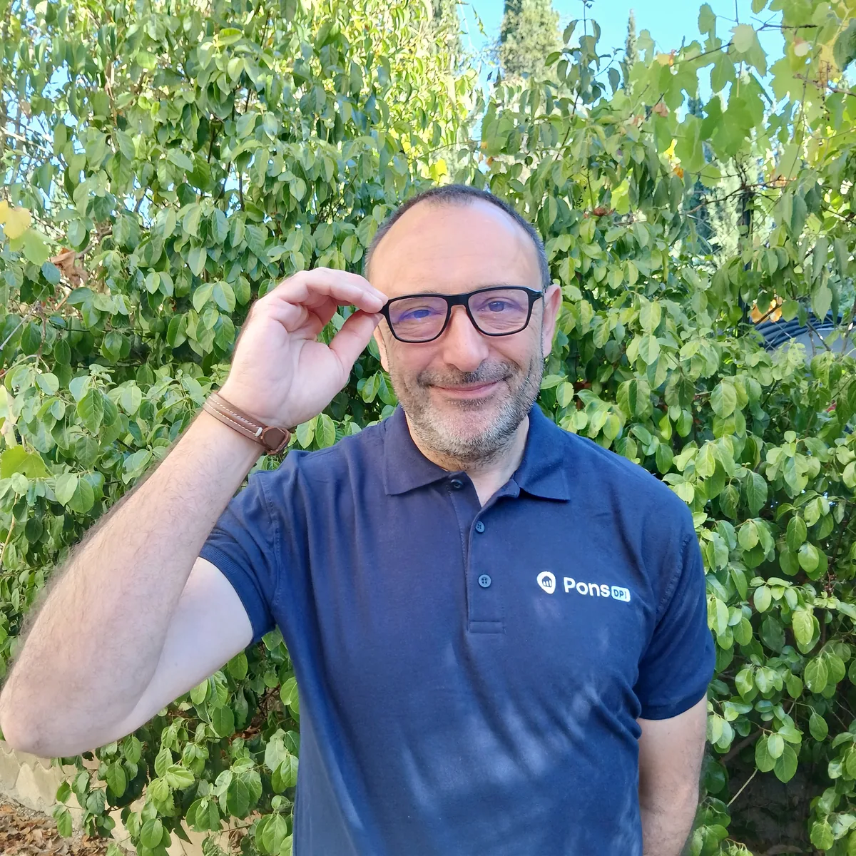 Guillaume Pons, diagnostiqueur immobilier à Montpellier, ajustant ses lunettes en souriant, en polo Pons DPI devant une haie